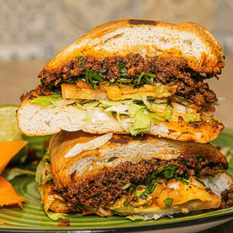 A large Mexican torta sandwich, cut in half, revealing seasoned meat, lettuce, and avocado on a green plate.