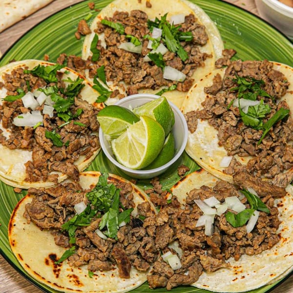 Five carne asada tacos with cilantro, onions, and lime wedges on a green plate.
